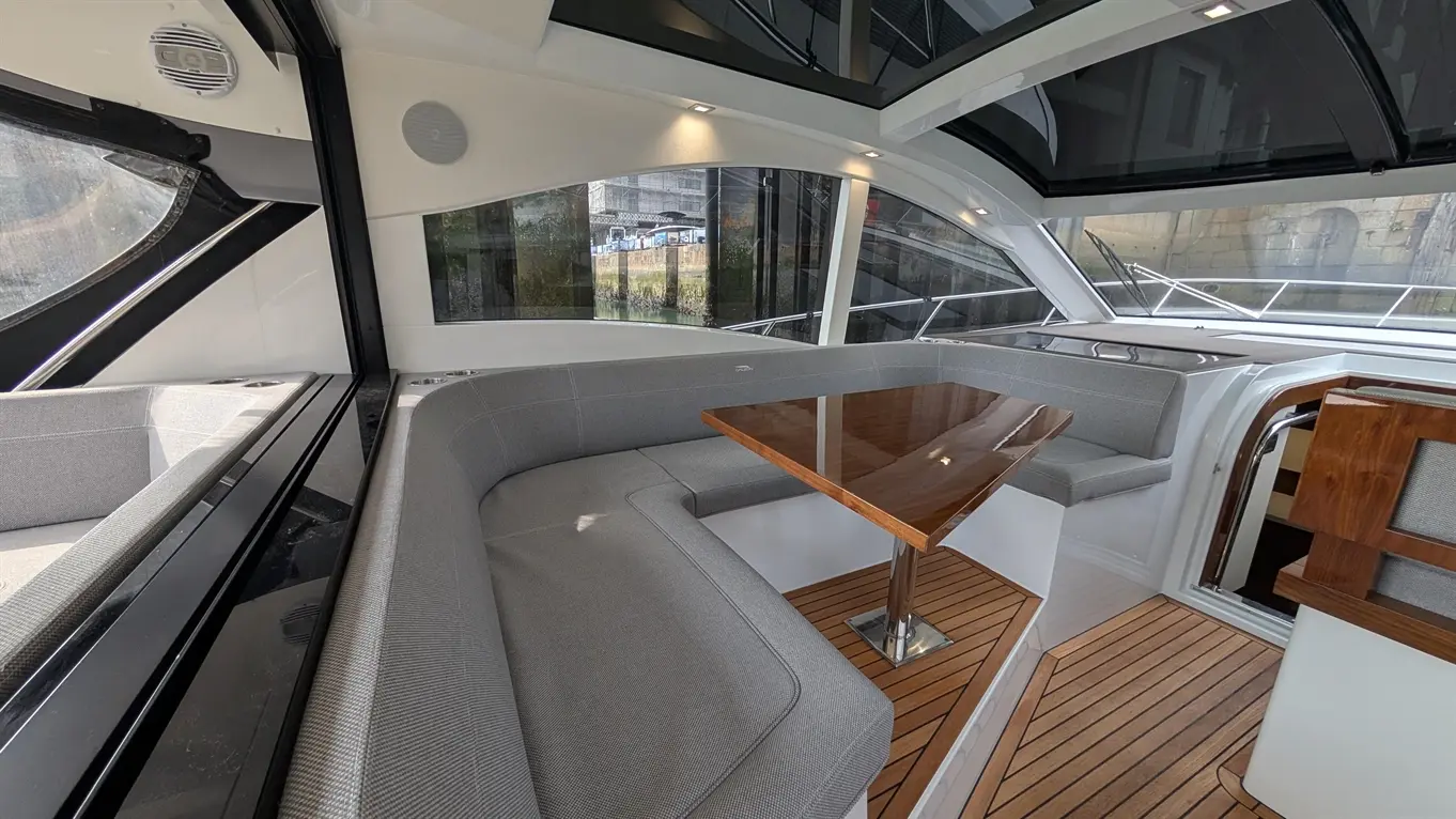 Luxurious yacht interior featuring grey seating, a polished wooden table, and large glass windows overlooking the marina.